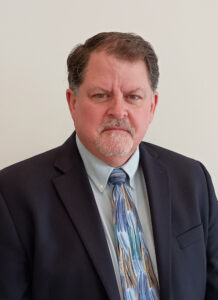 Portrait of a middle-aged man with short brown and gray hair and a neatly trimmed goatee, wearing a dark suit jacket, light blue dress shirt, and a patterned blue and tan tie, standing against a plain light background.