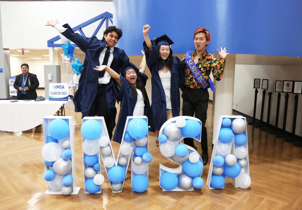 Students leap in graduation gowns in front of large bubble letters that spell out IMSA